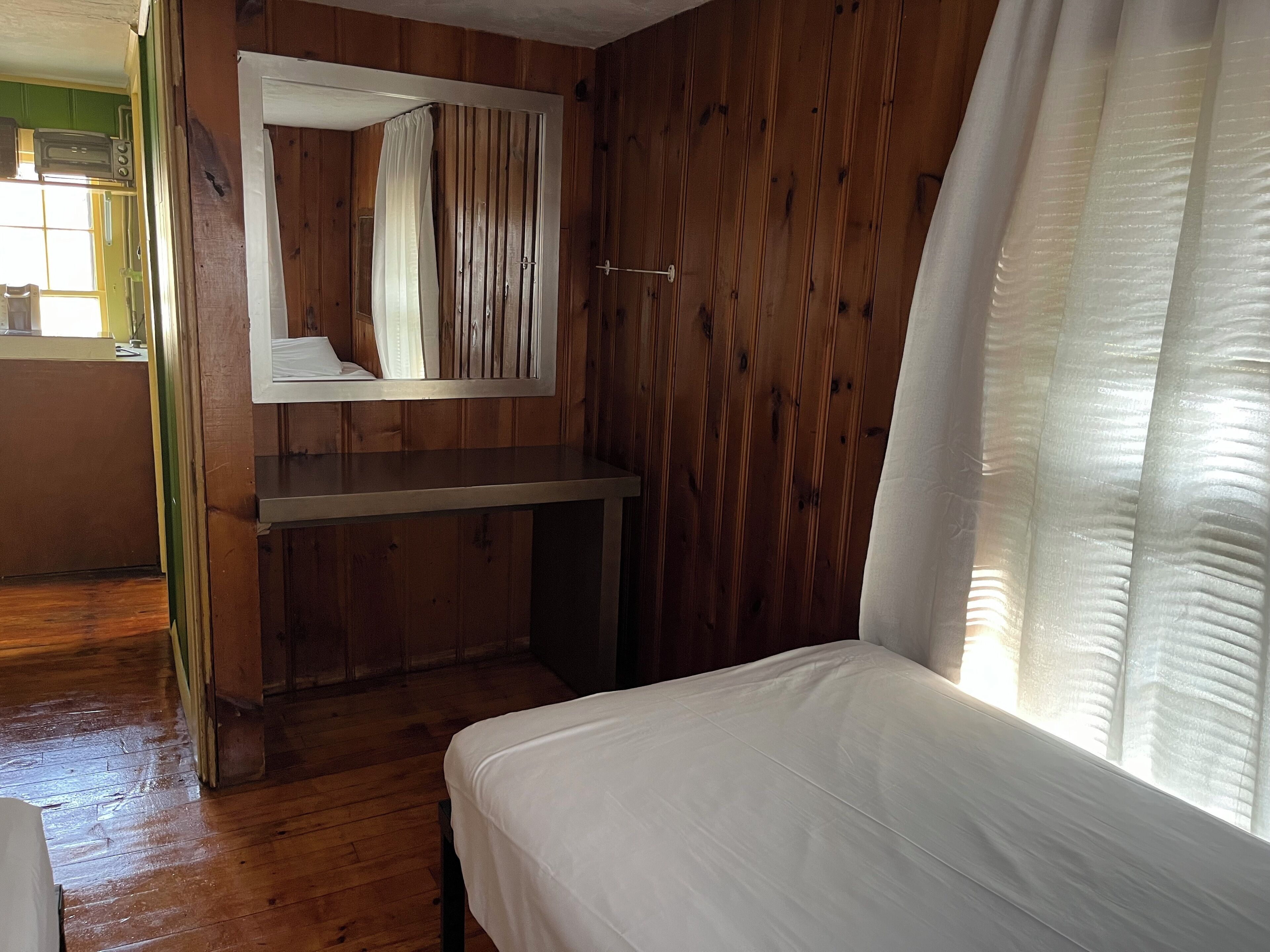 Una habitación de madera con una cama, un espejo y un ventanal con cortinas.