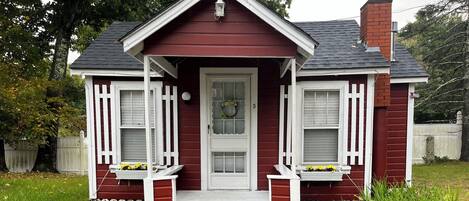 A small, single-story house with a red exterior and white trim, featuring a front porch with two windows and a door.