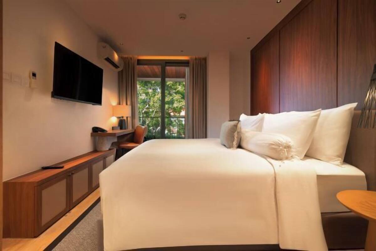 A hotel room with a large bed, a TV mounted on the wall, a desk with a chair, and a window with a view of greenery.