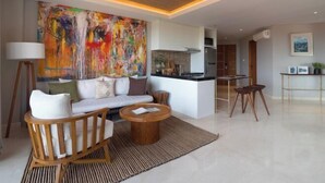 A modern living room with a sofa, coffee table, and chairs, a large abstract painting on the wall, and a kitchen area in the background. - Beachwalk Residence (Bali)