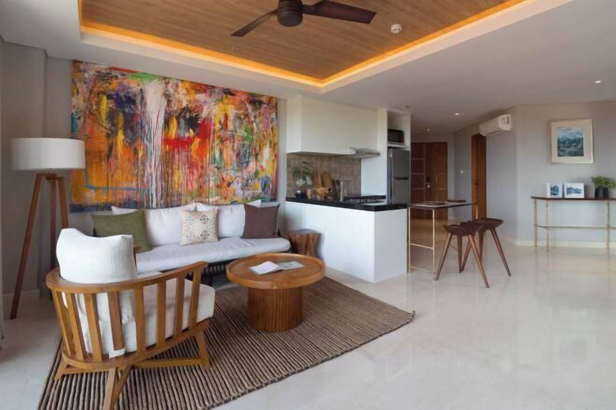 A modern living room with a sofa, coffee table, and chairs, a large abstract painting on the wall, and a kitchen area in the background.