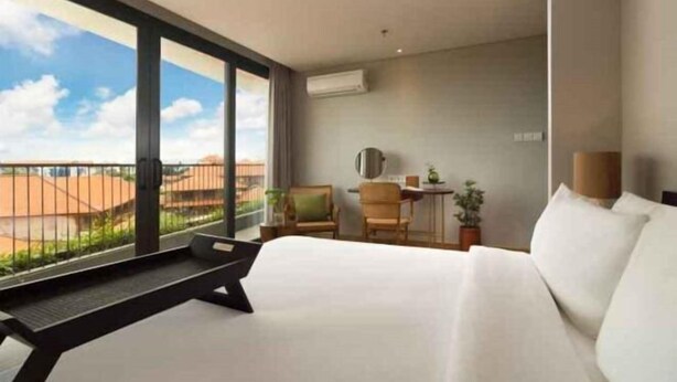A hotel room with a large bed, a desk, two chairs, a small table, and a view of the landscape through large windows. - Beachwalk Residence (Bali)
