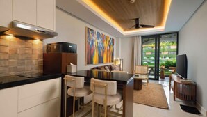 A modern kitchen with a black countertop, white cabinets, and a large painting. There is a dining area with a sofa and chairs, and a view of a garden through a glass door. - Beachwalk Residence (Bali)