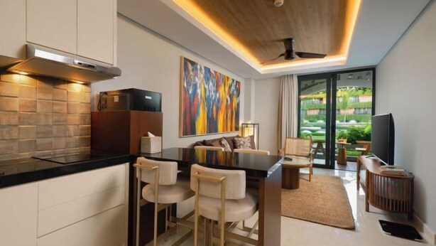 Una cocina moderna con encimera negra, armarios blancos y un cuadro grande. Hay una zona de comedor con sofá y sillas, y vista a un jardín a través de una puerta de cristal.