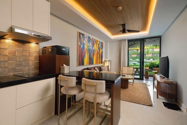 A modern kitchen with a black countertop, white cabinets, and a large painting. There is a dining area with a sofa and chairs, and a view of a garden through a glass door. - Beachwalk Residence (Bali)