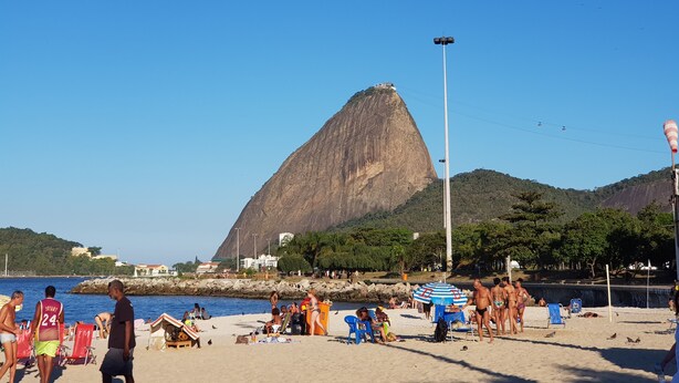 Une plage avec des gens, des chaises et des parasols. Une grande montagne se dresse à l’arrière-plan.