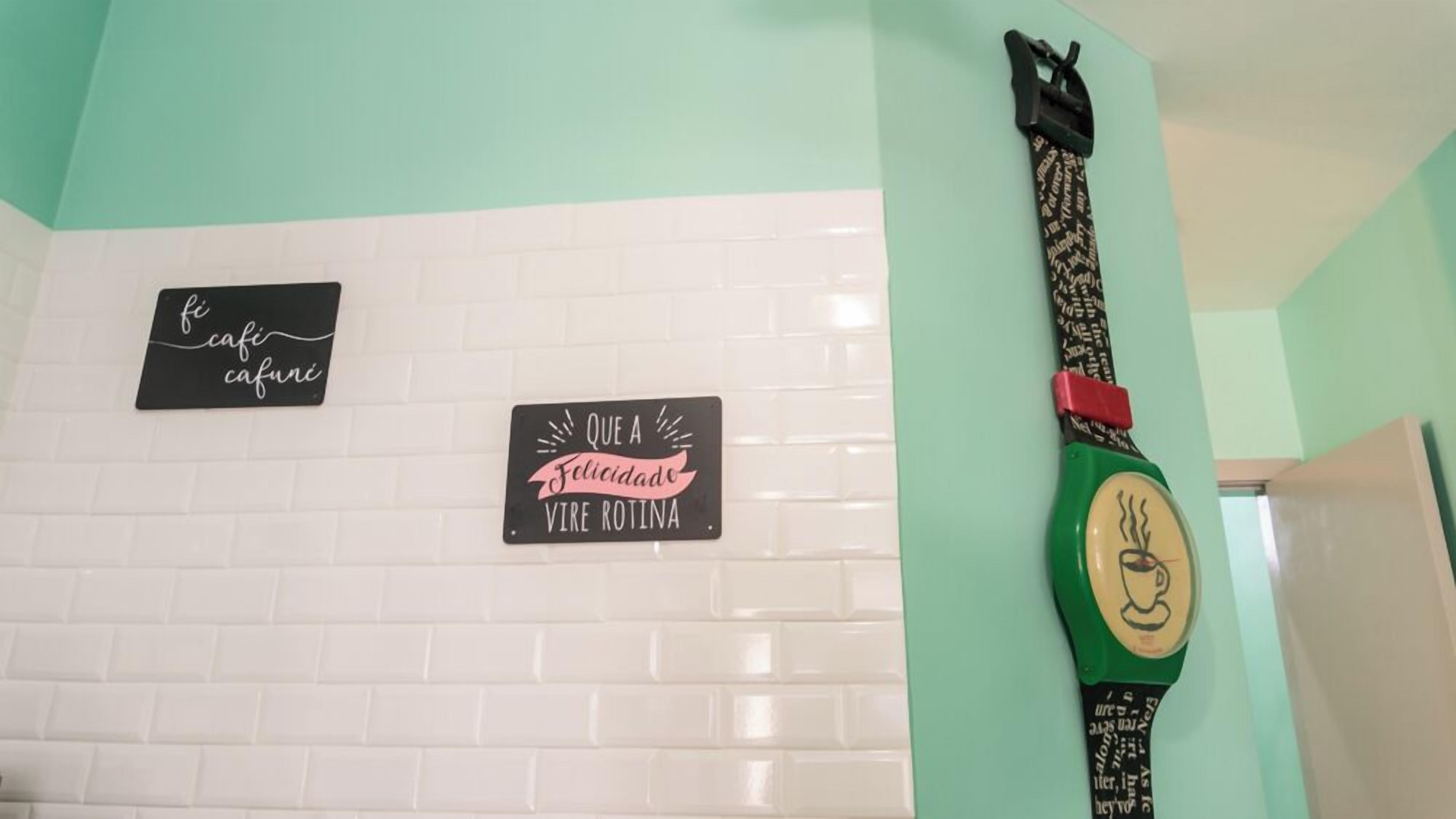 A wall with two signs and a clock. The signs read "fi cafe cafune" and "Que A Ficidado Vire Rotina." The clock has a green strap and a face with a coffee cup design.