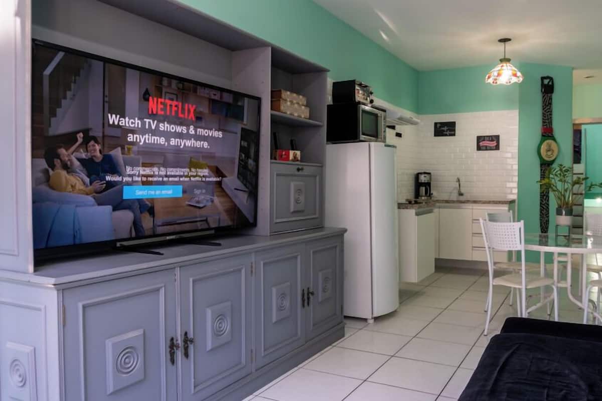 Een woonkamer met een flatscreen-tv waarop een Netflix-advertentie wordt weergegeven, een keuken met witte kasten, een eettafel met stoelen en een zwarte bank.