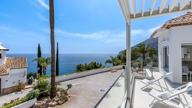 A balcony with a view of the sea, white lounge chairs, and a building with a tiled roof.