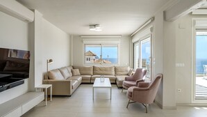 A modern living room with a sectional sofa, a coffee table, and a TV. There are large windows offering a view of the sea.