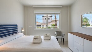 A bedroom with a bed, a chair, a desk, and a window with a view of a building and greenery.