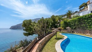 A swimming pool with a curved edge, surrounded by a stone wall and a grassy area. There are trees and a view of the sea and a mountain in the background.