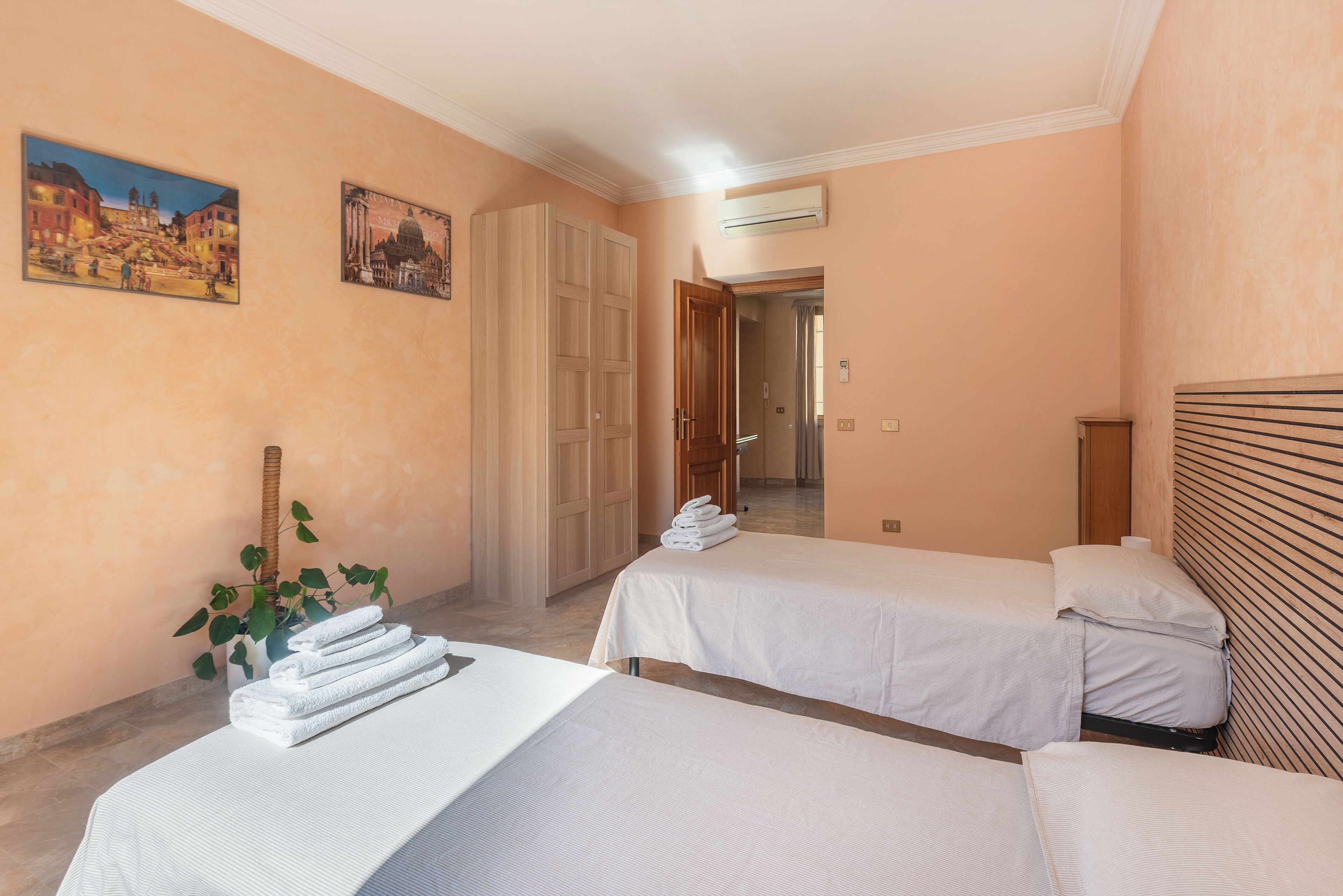 A hotel room with two beds, a potted plant, and a hallway leading to another room.