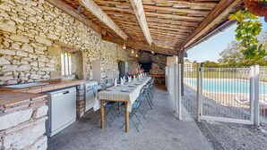 An outdoor dining area with stone walls, a wooden pergola, a pool, and a grill. - Presbytère de Pompiac (Castillonnès)