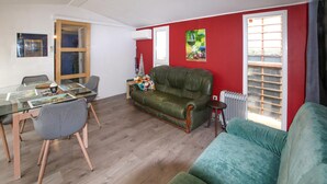 Ein Wohnzimmer mit einem grünen Sofa, einem Esstisch mit Stühlen, einer roten Akzentwand und einem Fenster mit Jalousien.