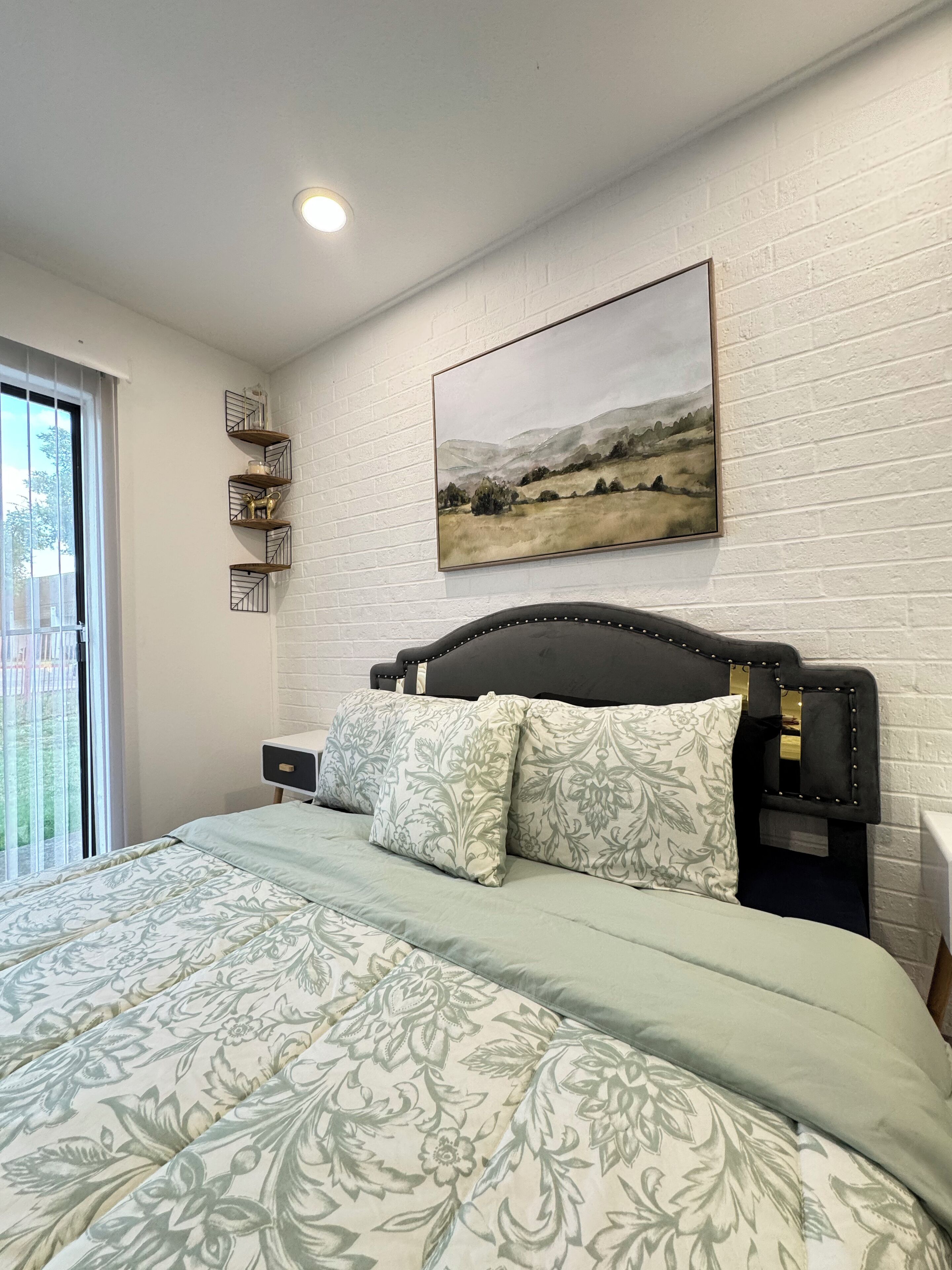 A bedroom with a bed, a nightstand, a wall-mounted shelf, and a painting.