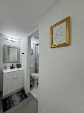 Une salle de bain avec un meuble-lavabo blanc, un miroir avec un cadre en marbre, une œuvre d’art encadrée en doré et un tapis à motifs noir et blanc.
