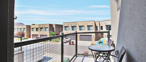 A balcony with a table and chairs, overlooking a street with parked cars and buildings.