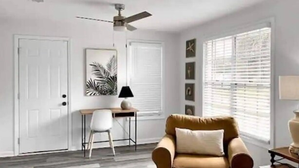 A living room with a beige armchair, a desk with a lamp, a ceiling fan, and a large window with blinds. - Tidy Apartment Near Hospital/PCC (Pensacola)