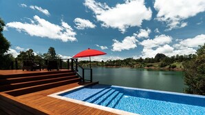 Una terraza de madera con piscina, una sombrilla roja y vistas a un lago y colinas.