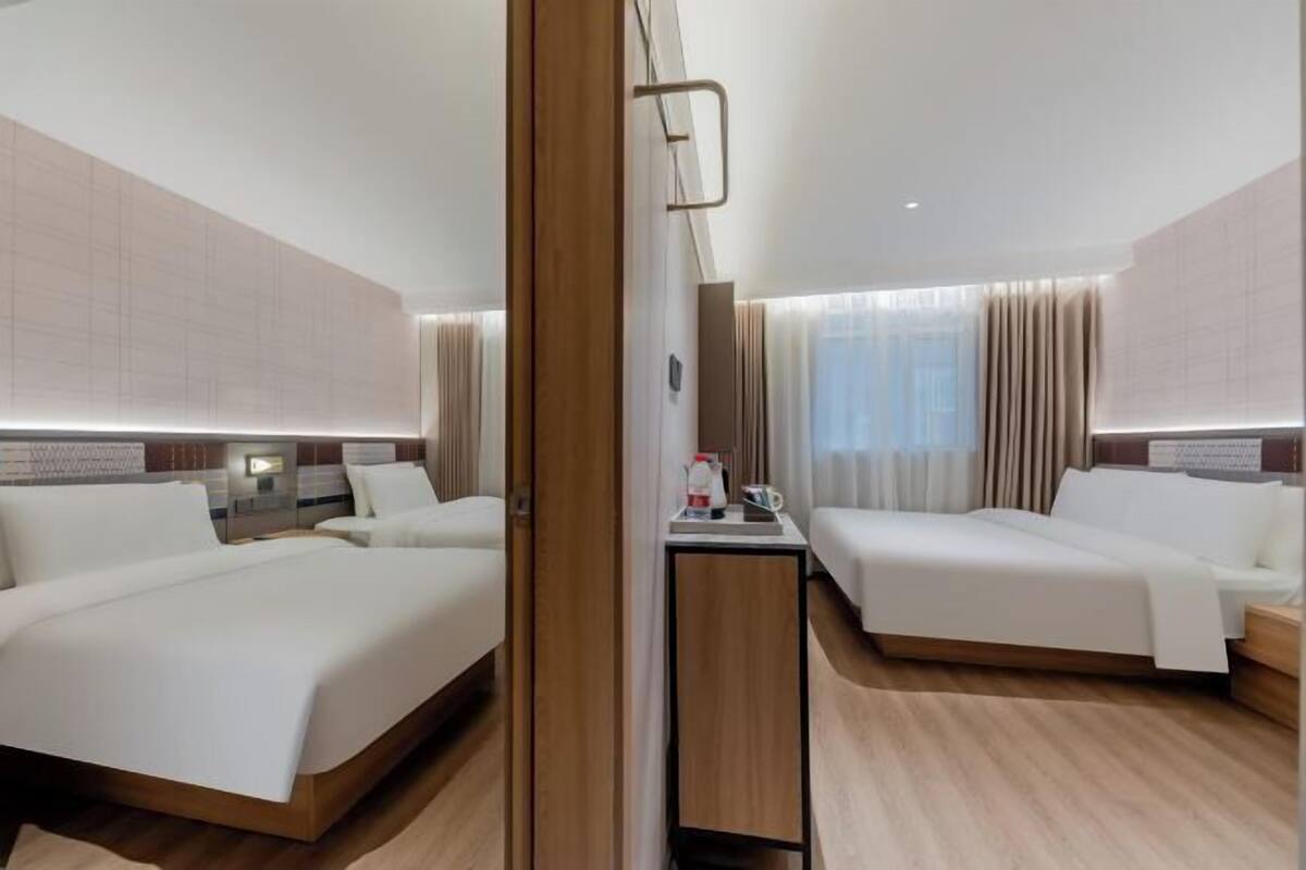 A hotel room with two beds, a bedside table, a mirror, and a window with curtains.