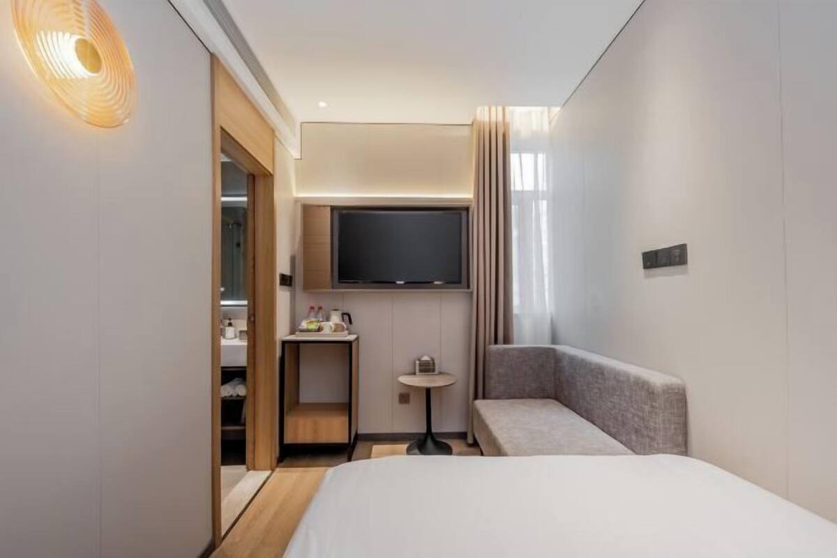 A modern hotel room with a bed, a seating area, a small table, a TV mounted on the wall, and a closet with a mirror.