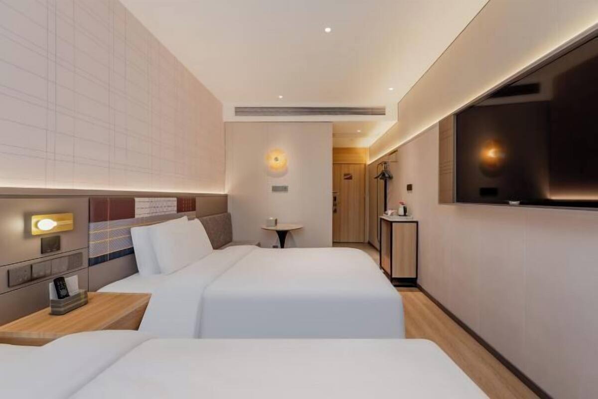 A hotel room with two beds, a small table, a bedside lamp, and a view of the hallway.