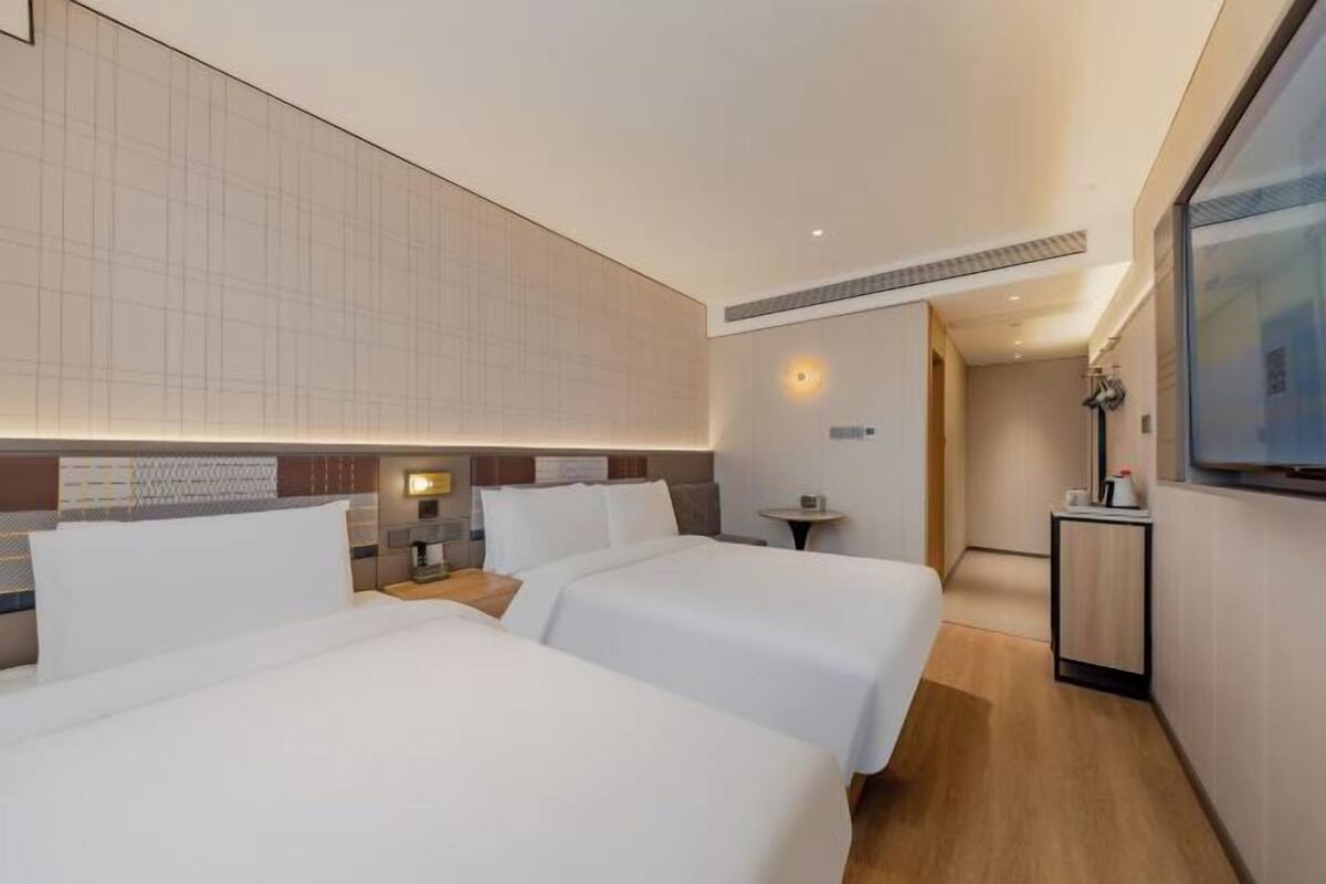 A hotel room with two beds, a small table, a TV, and a wall-mounted light.