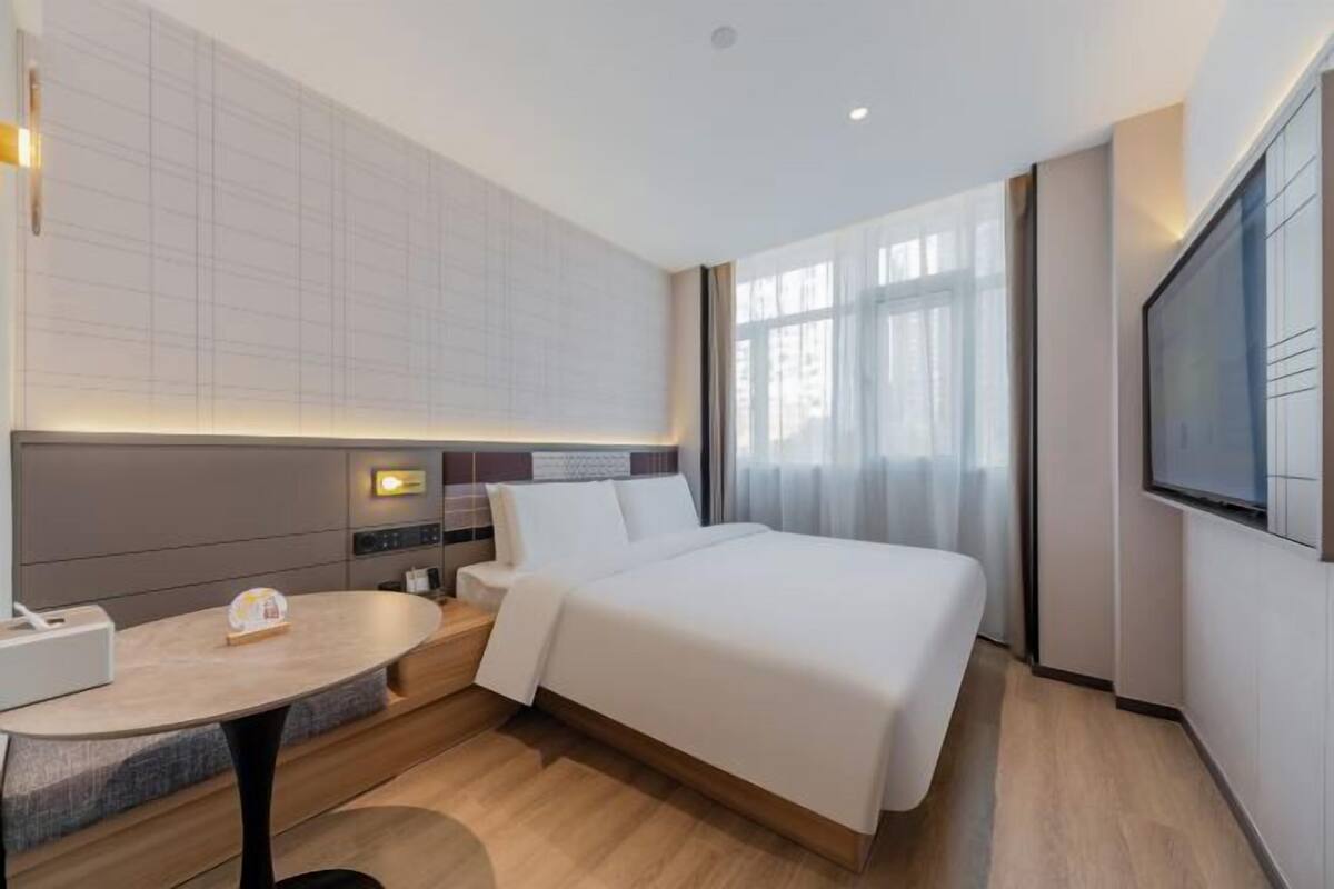 A modern hotel room with a large bed, a bedside table, a desk, and a TV mounted on the wall.