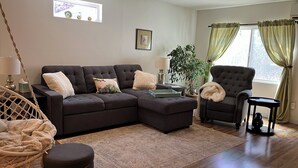 A modern living room with a sectional sofa, a round ottoman, a hanging chair, a side table with a lamp, and a potted plant. - Charming cottage in scenic Seattle with AC, WiFi, and all the comforts of home. (Seattle)