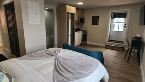A bedroom with a bed, a desk, a chair, a kitchenette, and a bathroom.