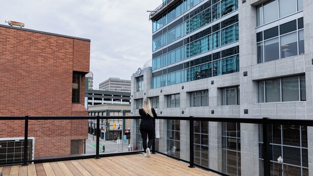 A wooden deck with a black railing, overlooking a modern building with large windows and a brick facade.