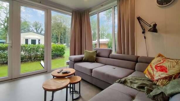 Ein modernes Wohnzimmer mit einem grauen Sofa, zwei kleinen runden Tischen und einem großen Fenster mit Blick auf einen Garten und ein Haus.