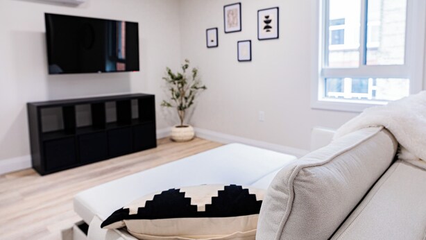 A modern living room with a flat-screen TV, a black entertainment unit, a white sofa, and a window with a view of buildings. - 1 Bedroom Unit with living space and full kitchen Prime Downtown location (Kitchener)