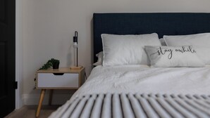 A neatly made bed with white bedding and two pillows, one with text. There is a bedside table with a lamp and a small plant. - 1 Bedroom Unit with living space and full kitchen Prime Downtown location (Kitchener)