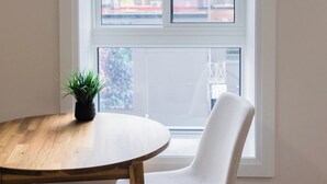 Une pièce avec une grande fenêtre, une table ronde en bois, une chaise blanche et une petite plante en pot.