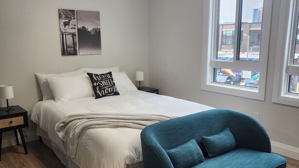 Une chambre à coucher avec un lit, un fauteuil bleu, une table de chevet, une lampe et une fenêtre donnant sur la ville.