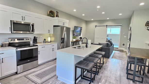 A modern kitchen with white cabinetry, stainless steel appliances, a central island with bar stools, and a dining area with a table and chairs. - RAINEY CREEK RETREAT (Swan Valley)