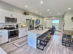 A modern kitchen with white cabinetry, stainless steel appliances, a central island with bar stools, and a dining area with a table and chairs. - RAINEY CREEK RETREAT (Swan Valley)
