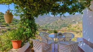 Terraza con sillas y mesa de celosía blanca, rodeada de vegetación y vistas a un paisaje montañoso.