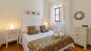 A bedroom with a bed, two bedside tables, a dresser, a window with curtains, and wall-mounted decor. - Casa Fuentezuelas Sayalonga by Ruralidays (Málaga)