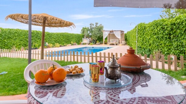 A table with oranges, a teapot, and colorful cups, set against a poolside view with a wooden fence and greenery. - Huerta Segura Antequera by Ruralidays (Málaga)