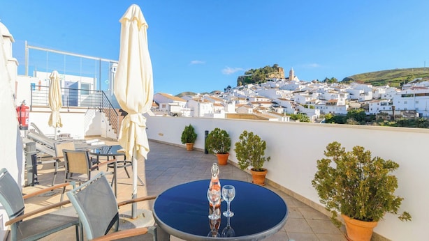 Une terrasse sur le toit, avec une table dressée pour deux personnes, des plantes en pot et une vue sur une ville aux murs blanchis à la chaux, avec une colline et une église.
