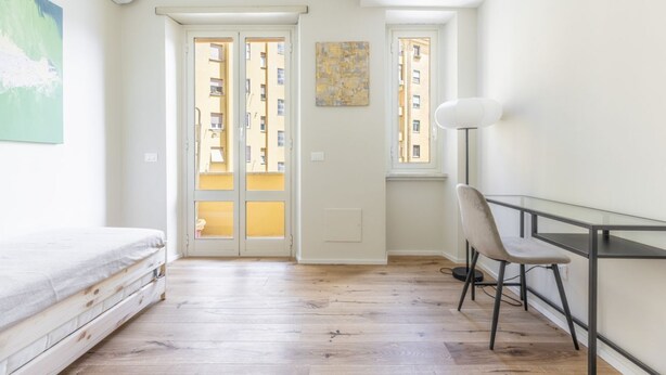 Une pièce comprenant un lit, un bureau, une chaise, une lampe sur pied et une fenêtre donnant sur des immeubles.