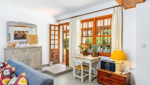 A cozy living room with a sofa, a wooden table, and a lamp. There is a window with curtains and a view of greenery outside.