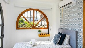 Un dormitorio con un gran ventanal de arco, una cama con ropa de cama estampada, una mesita de noche con lámpara y una pared decorada.