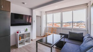 Ett modernt vardagsrum med en platt-TV, ett soffbord i glas, en grå soffa och utsikt över en stadssiluett genom stora fönster.