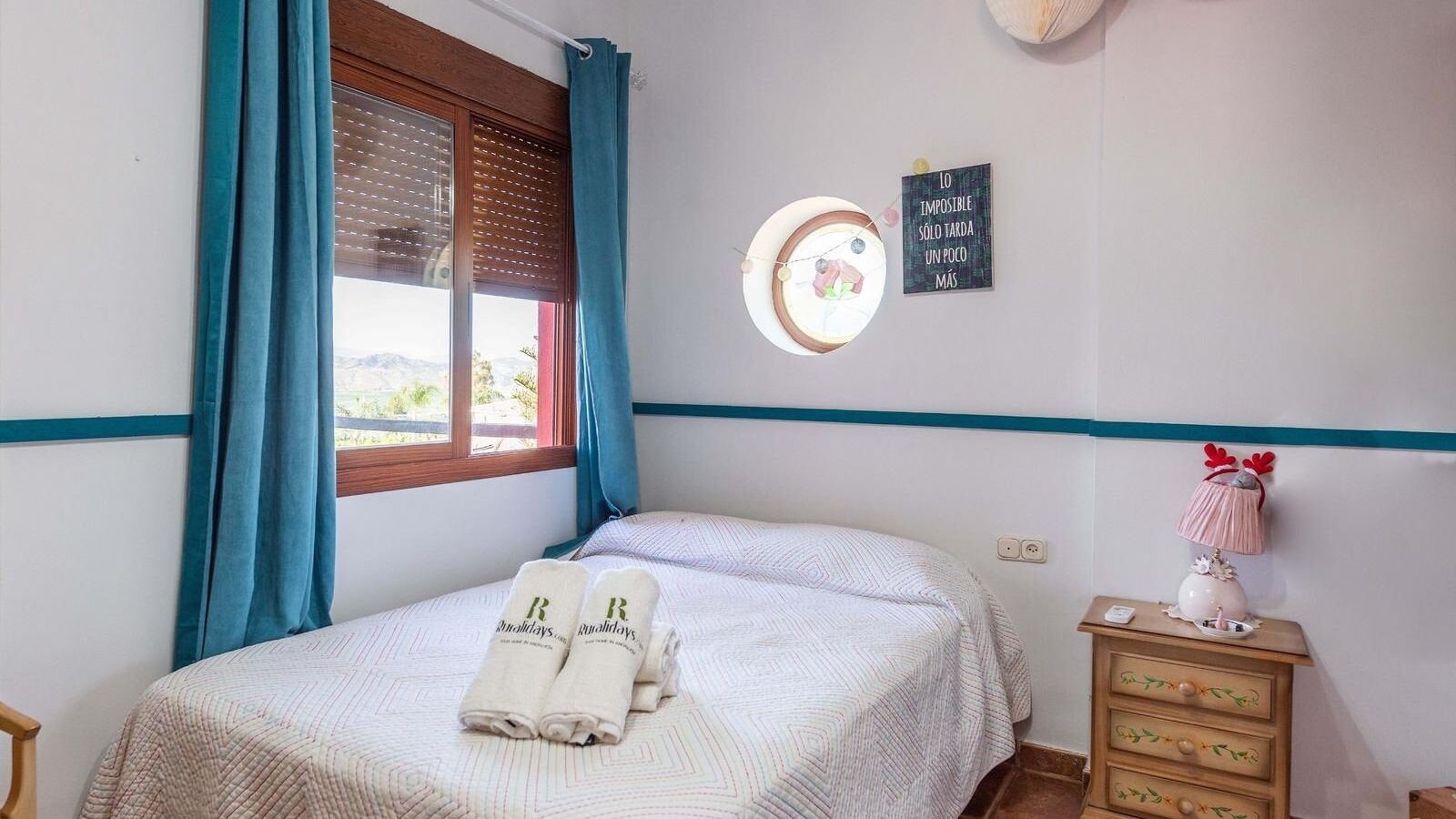 Cama bem arrumada com colcha listrada branca e clara, duas toalhas enroladas com um logotipo, mesa de cabeceira com abajur e objeto decorativo, e parede com janela circular e frase motivacional.