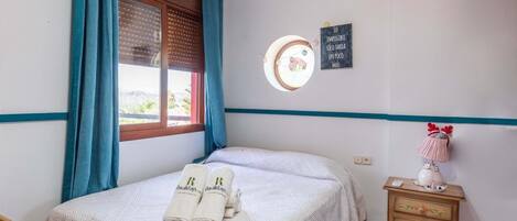 Cama bem arrumada com colcha listrada branca e clara, duas toalhas enroladas com um logotipo, mesa de cabeceira com abajur e objeto decorativo, e parede com janela circular e frase motivacional.