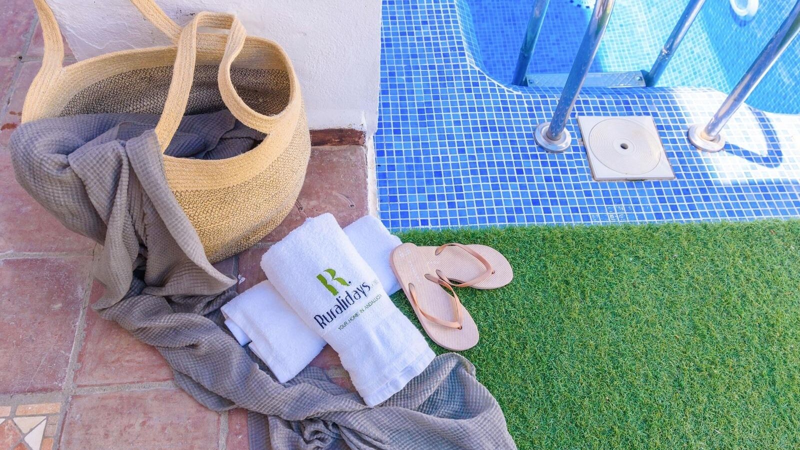 Area piscina con pavimento in piastrelle blu, un cesto in vimini, un asciugamano e ciabatte, e un asciugamano bianco con un logo.